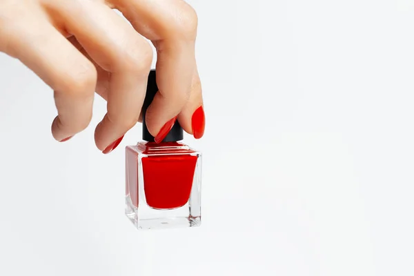 Close-up of female hand with red manicure holding a glitter nail polish of red color on white background.
