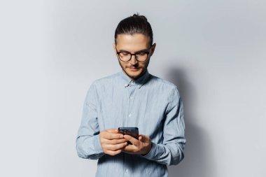 Beyaz arka planda akıllı telefon kullanan, mavi gömlek ve gözlük takan genç bir adamın stüdyo portresi..