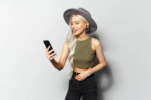 Studio portrait of cute happy blonde girl while dancing and listen the music via wireless earbuds, holding smartphone in hand, wearing grey hat on white background.