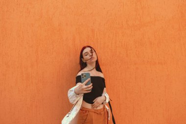 Kızıl saçlı güzel bir kız telefonuyla duvara selfie çekiyor.