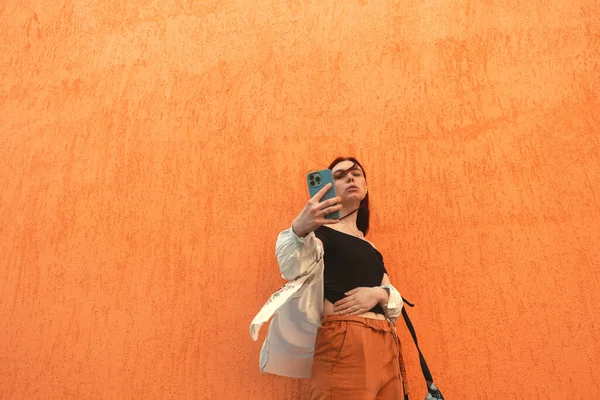 Kızıl saçlı güzel bir kız telefonuyla duvara selfie çekiyor.