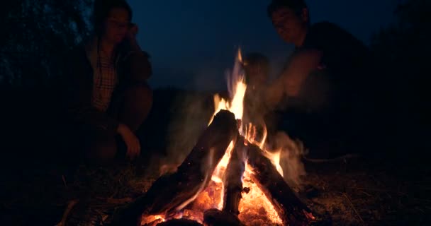 Family Sit near Campfire Watching on Fire Flame. Tourist Father Mother Child Rest on Camping Site Near Lake in Field Plains. Rural Country Countryside Scene. Summer Evening Twilight Dusk Night 4K