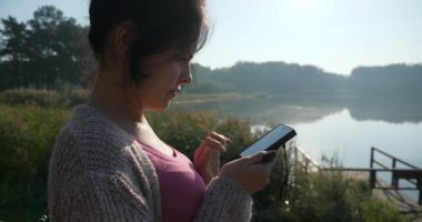 Genç Pretty Woman, Şehir Parkı 'ndaki Güzel Doğa' da Dinlenen Akıllı Telefon Dokunmatik Mesajlar Kullanıyor. Yaz Sabahı Güneşli Gün 4K