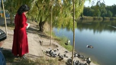 Kırmızı Uzun Paltolu Güzel Kadın Şehir Parkı 'ndaki Göl Göleti yakınlarında güvercinleri besliyor. Güneşli bir gün. 2x Yavaş çekim 60fps 4K