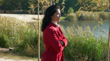 Kırmızı paltolu güzel kadın göl kenarında yürüyor. Park 'ta dinleniyor. Ciddi Düşünceli Kadın Duygu İfadesi. Güneşli bir sonbahar günü. Yavaş çekim 4K
