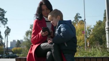 Anne ve Oğul Medya 'yı telefonda izleyin. İnternette Sörf Yapan Kadın Akıllı Telefon Kullanıyor. Annem ve Kid şehir parkında dinleniyor. Sonbahar Günü. 2x Yavaş çekim 60fps 4K