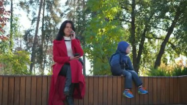Meşgul genç anne telefonda konuşuyor. İş kadını, Parkta Çocuğa Bakıcılık Yapan Muhabbet Kadını İçin Akıllı Telefon Kullan. Dişi Kırmızı Uzun Paltolu. 2x Yavaş çekim 60fps 4K