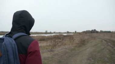 Nehir doğası yakınlarındaki kırsal alanda düşünceli bir adam yürüyüş yapıyor. Erkek Turist Maceracı Gezgin Sonbahar Bulutlu Günü. 2x Yavaş çekim 60fps 4K