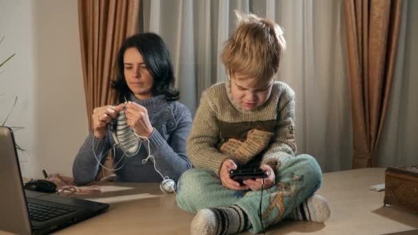 儿童男孩使用的是观看智能手机互联网社交媒体 母亲针织针织针织在窗前 寒冬季节 2X慢动作60Fps — 图库视频影像