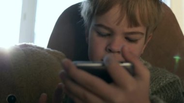 Lonely Boy with Teddy Bear Watching Smartphone Internet Social Media. Kid  Use Modern Phone Gadget. Child Wearing Sweater Sit near Window. Cold Winter Season. 2x Slow motion 60 fps 4K