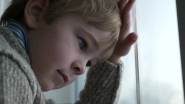 Üzgün küçük çocuk pencerenin yanında duruyor. Düşünceli yalnız çocuk, bekleyin bayan. Çocuk süveteri giy. Kış Mevsimi Günü. 2x Yavaş çekim 60fps 4K
