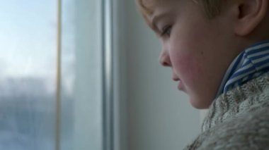 Üzgün küçük çocuk pencerenin yanında duruyor. Düşünceli yalnız çocuk, bekleyin bayan. Çocuk süveteri giy. Kış Mevsimi Günü. 2x Yavaş çekim 60fps 4K