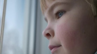 Üzgün küçük çocuk pencerenin yanında duruyor. Düşünceli yalnız çocuk, bekleyin bayan. Çocuk süveteri giy. Kış Mevsimi Günü. 2x Yavaş çekim 60fps 4K