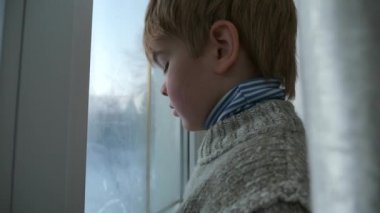 Üzgün küçük çocuk pencerenin yanında duruyor. Düşünceli yalnız çocuk, bekleyin bayan. Çocuk süveteri giy. Kış Mevsimi Günü. 2x Yavaş çekim 60fps 4K