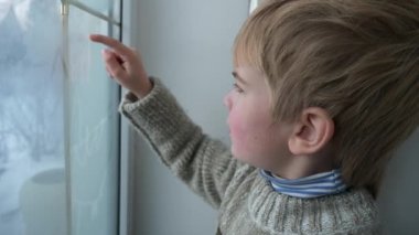 Sevimli küçük çocuk ıslak camlara parmak çiziyor. Çocuk süveter giyiyor. Kış Mevsimi Günü. 2x Yavaş çekim 60fps 4K