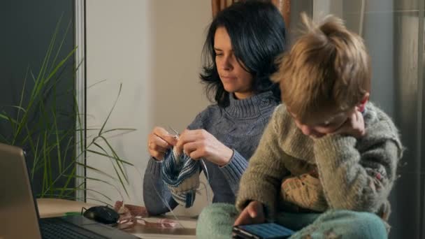 子供の男の子は スマートフォンのインターネットソーシャルメディアを見て使用します 母は窓の近くで編み物の針で編む 寒い冬の季節 2倍スローモーション60Fps — ストック動画