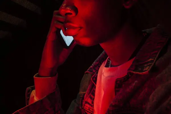 身穿斜纹棉布夹克的非裔美国人沉着严肃的一面 穿着霓虹灯在手机上黑色背景的神秘红光 — 图库照片