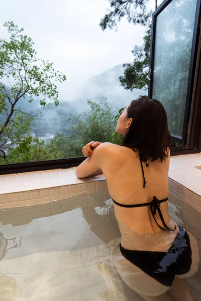 山と緑の木々を眺めながら 窓のそばの温泉街にある日本の温泉に座っている水着姿のリラックスした若い民族女性 — ストック写真
