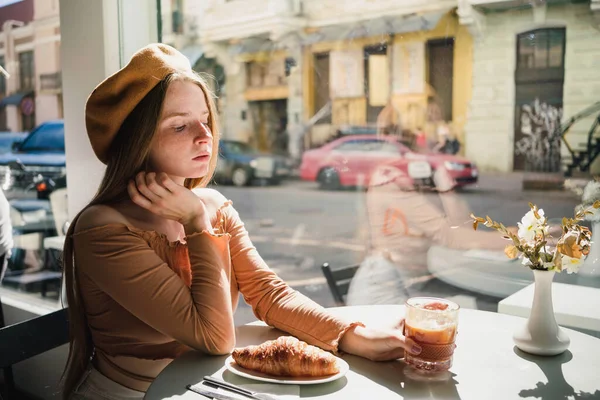 ベレー帽のフランス人女性はコーヒーの香り高いガラスと焼きたてのクロワッサンとカフェのテーブルに座っています — ストック写真