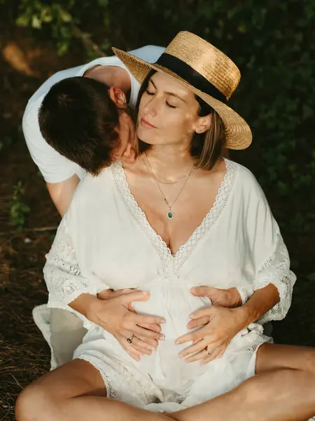 男抱擁とキス首の妊娠中の女性で目を閉じて後ろから田舎の牧草地に座って日没 — ストック写真