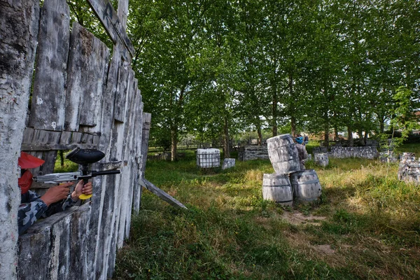 ゲーム中に木製の樽の後ろに隠れて敵を目指してペイントボール銃を持つ匿名の男性シューティングゲーム — ストック写真
