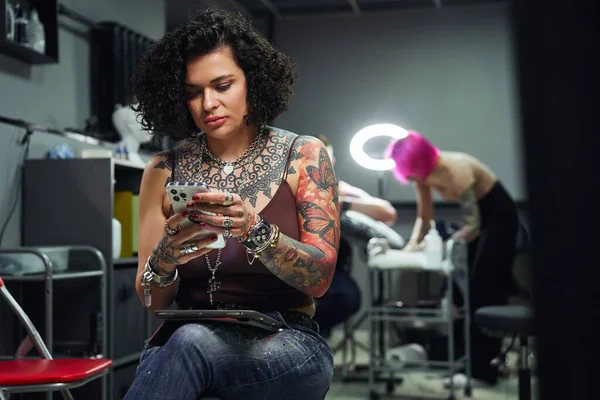身穿休闲装 头戴纹身的自信成年女性坐在智能手机上冲浪时 靠近纹身师的明亮纹身沙龙为客人做纹身 — 图库照片
