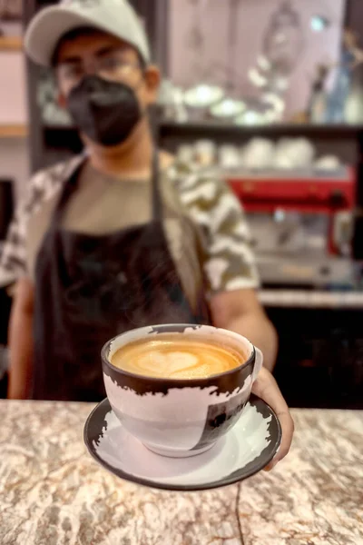 カフェテリアでカメラを見ながら ラテアートとアロマコーヒーのカップを提供顔マスクのエスニック男 — ストック写真