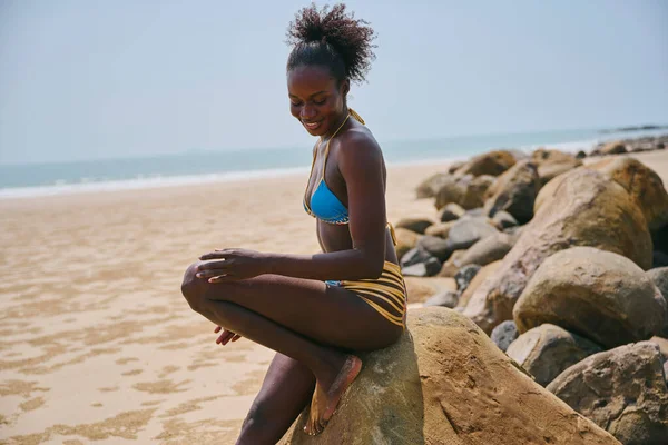 サイドビューのコンテンツ若いアフリカ系アメリカ人女性の水着岩の上に座って砂の海の海岸を見下ろしながら — ストック写真