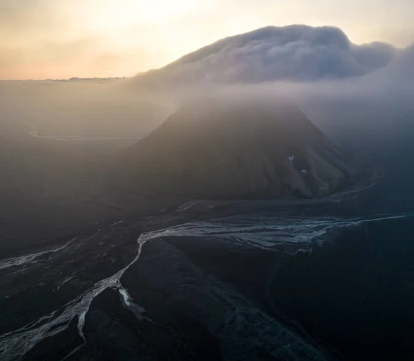 険しい表面を霧で覆われた火山山は 夕方には雲一つない空に対して自然の流れ川の近くに位置しています — ストック写真