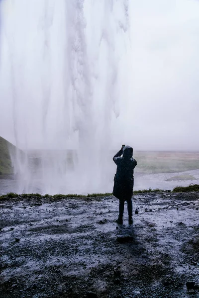 在冰岛旅行期间 穿着保暖外套和帽衫的难以辨认的游客在智能手机上拍下了色彩艳丽的塞亚尔和福斯瀑布的背影 — 图库照片