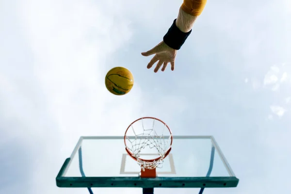 青い空に対して公共のスポーツの地面にゲームをプレイしながら輪にバスケットボールボールを投げる作物の匿名の人の下から — ストック写真