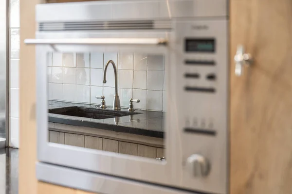 金属製の蛇口とフラットの現代的なキッチンでオーブンに内蔵されたガラスのドアに反映されたアンマウントシンク — ストック写真