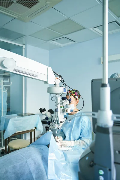 入院中の認識できない患者の目を操作しながら顕微鏡を通して見て外科制服や滅菌マスクの注目すべき女性医師 — ストック写真