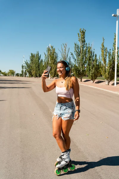 ポジティブな女性スケーターでローラーブレードとヘッドフォン取ります自己撮影上の携帯電話で晴れた日に夏に都市で — ストック写真