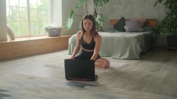 Μια νεαρή γυναίκα ασχολείται με online προπονήσεις στο σπίτι. Το λάπτοπ είναι στο πάτωμα και η κοπέλα πάει μόνη της για σπορ.. — Αρχείο Βίντεο