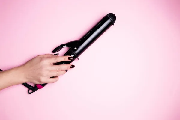 Female hand holds a hair curler in black on a pink isolated background close-up. — Stock Photo, Image