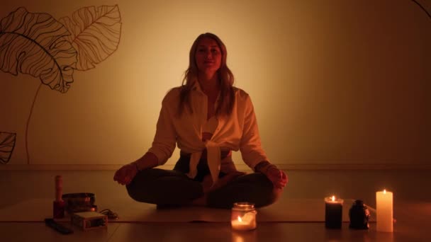 Žena sedí v lotosové pozici v šeru a na koberci hoří svíčky. Trénink jógy a totální relaxace. — Stock video