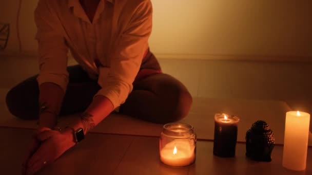 Una mujer se sienta en una posición de loto en el crepúsculo con velas ardiendo en la alfombra. Práctica de yoga y relajación total. — Vídeo de stock