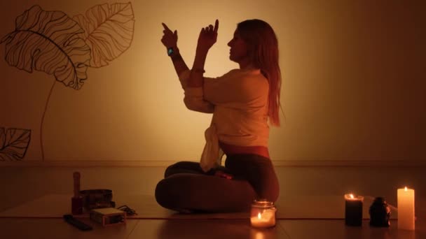 Eine Frau sitzt in einer Lotusposition in der Dämmerung, auf dem Teppich brennen Kerzen. Yoga-Praxis und totale Entspannung. — Stockvideo
