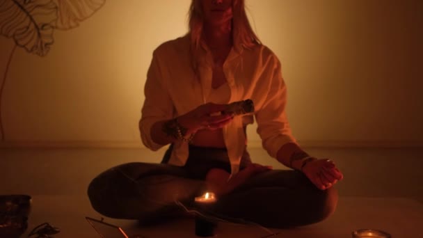 Девушка сидит на ковре и зажигает благовония для йоги и ароматерапии в комнате со свечами. — стоковое видео