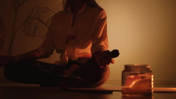 Een meisje zit op een tapijt en licht wierook voor yoga en aromatherapie in een kaarskamer. — Stockvideo