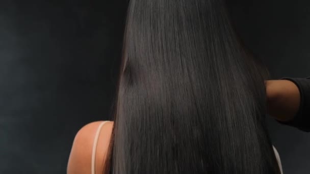 Nahaufnahme einer Brünetten mit luxuriösen Haaren auf dunklem Hintergrund. Das Mädchen steht mit dem Rücken und entwickelt die Haare. — Stockvideo