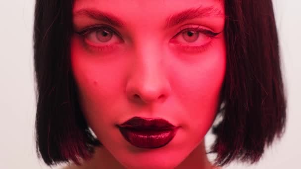 Retrato de una niña en luz roja con labios negros y una peluca sobre un fondo blanco en el estudio. Mujer en luz creativa de neón. — Vídeo de stock