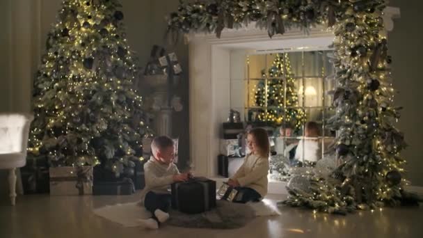 Szczęśliwe dzieci otwierają prezent pod choinką. Dom jest ozdobiony lampkami Nowego Roku. — Wideo stockowe