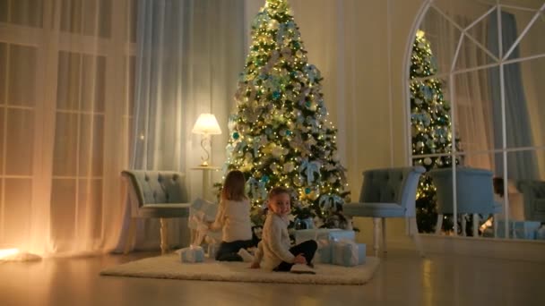 Happy children open a gift under the Christmas tree. The house is decorated with New Years lights. — Stock Video