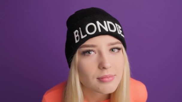 En vacker blond flicka tittar misstänksamt på kameran på en lila bakgrund. — Stockvideo
