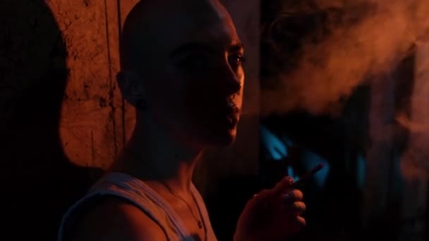 Portrait of a bald girl with tunnels in her ears and a cigarette close-up. Gloomy room in neon light. The girl smokes and blows out smoke. — Stock Video