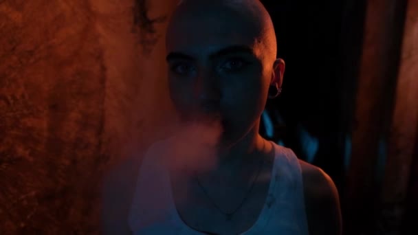 Porträtt av en skallig flicka med tunnlar i öronen och en cigarett närbild. Gloomy rum i neonljus. Flickan röker och blåser ut rök. — Stockvideo