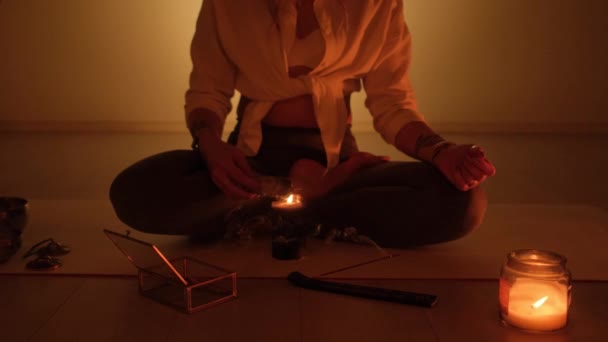 Ένα κορίτσι κάθεται σε ένα χαλί και ανάβει θυμίαμα για γιόγκα και αρωματοθεραπεία σε ένα δωμάτιο με κεριά. — Αρχείο Βίντεο