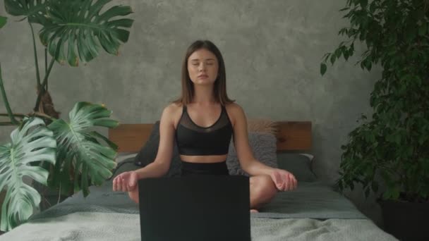 Das Mädchen praktiziert Yoga online mit einem Laptop zu Hause auf dem Bett. Entspannte Namaste-Pose. — Stockvideo
